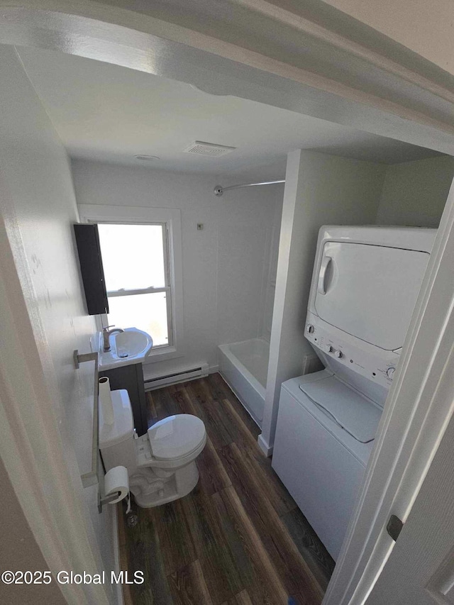 bathroom featuring stacked washer and dryer, toilet, a sink, a baseboard heating unit, and wood finished floors