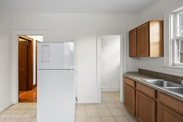 kitchen with a sink, freestanding refrigerator, brown cabinetry, light tile patterned floors, and baseboards