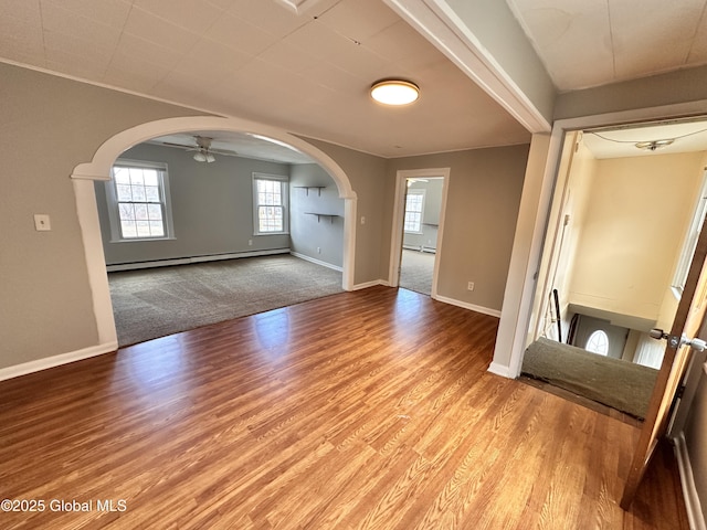 empty room with arched walkways, light wood finished floors, a ceiling fan, and a baseboard radiator