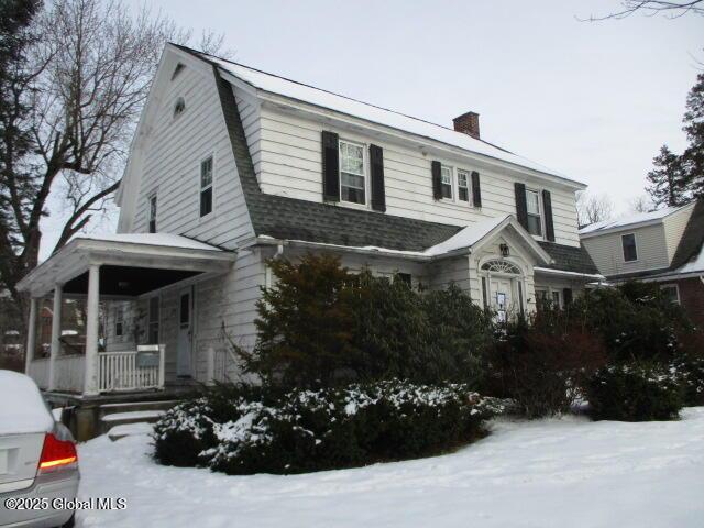 dutch colonial with a gambrel roof, roof with shingles, covered porch, and a chimney