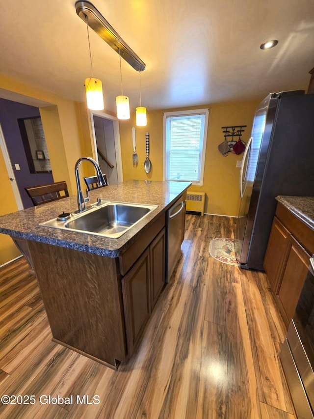 kitchen featuring radiator, an island with sink, dark wood-style floors, stainless steel appliances, and a sink