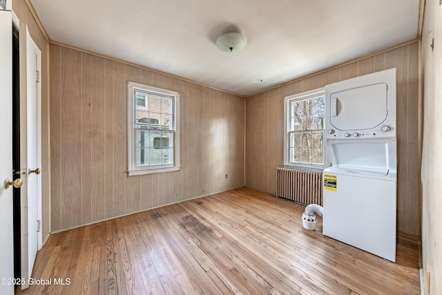 unfurnished room featuring wooden walls, stacked washing maching and dryer, radiator heating unit, and hardwood / wood-style floors