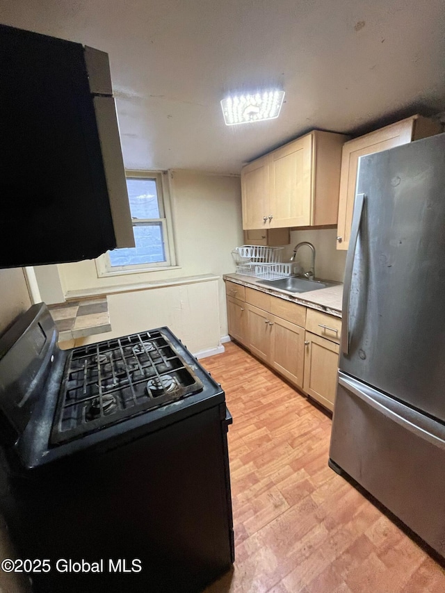 kitchen featuring black gas stove, light brown cabinetry, freestanding refrigerator, light wood-style floors, and a sink