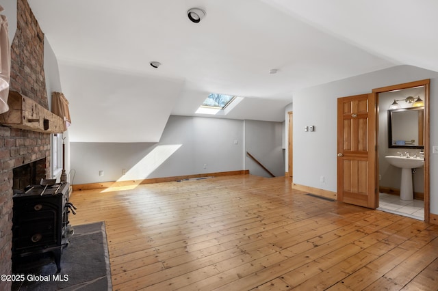 unfurnished living room with baseboards, vaulted ceiling with skylight, a wood stove, light wood-style floors, and a sink