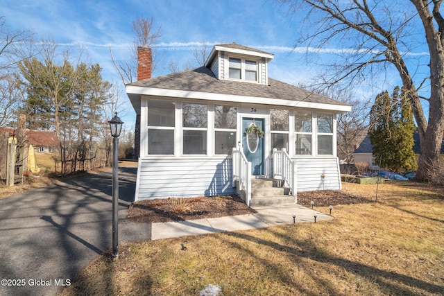 bungalow-style home with a front lawn, a chimney, a sunroom, and a shingled roof