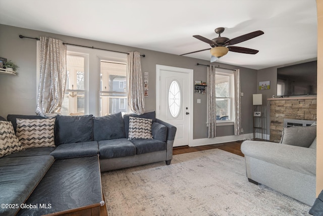 living area featuring a stone fireplace, wood finished floors, a ceiling fan, and baseboards