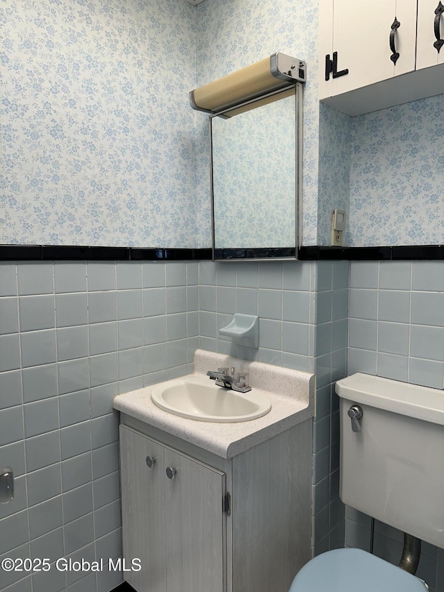 half bathroom with vanity, a wainscoted wall, wallpapered walls, tile walls, and toilet