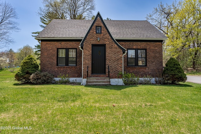 english style home with brick siding, roof with shingles, entry steps, and a front lawn