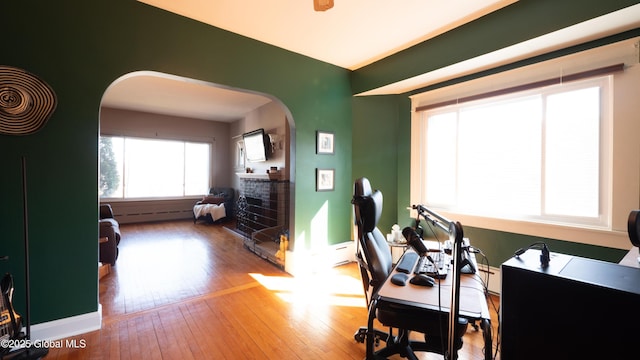 office area with hardwood / wood-style floors, a fireplace, baseboards, a baseboard heating unit, and arched walkways