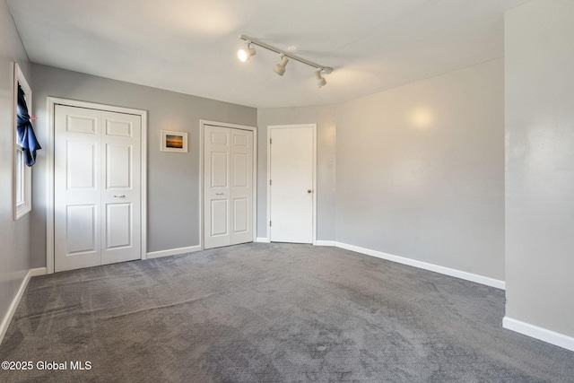 unfurnished bedroom with two closets, dark carpet, baseboards, and track lighting