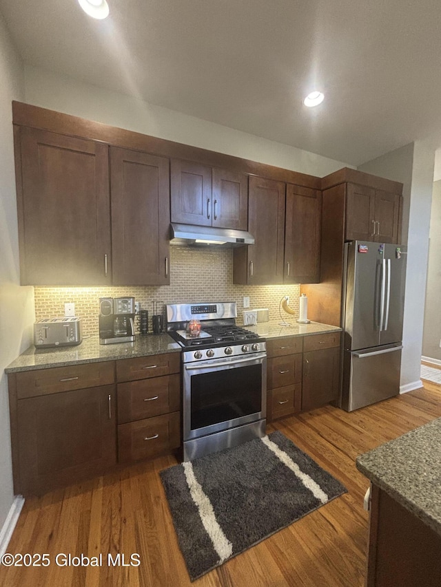 kitchen with light stone counters, wood finished floors, under cabinet range hood, appliances with stainless steel finishes, and tasteful backsplash