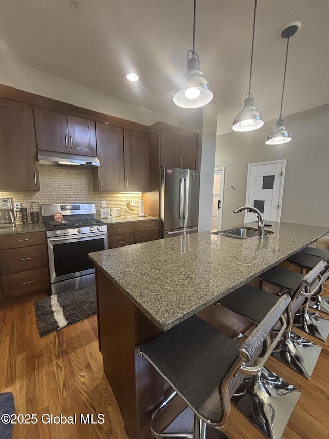 kitchen with wood finished floors, a sink, stainless steel appliances, under cabinet range hood, and a kitchen breakfast bar