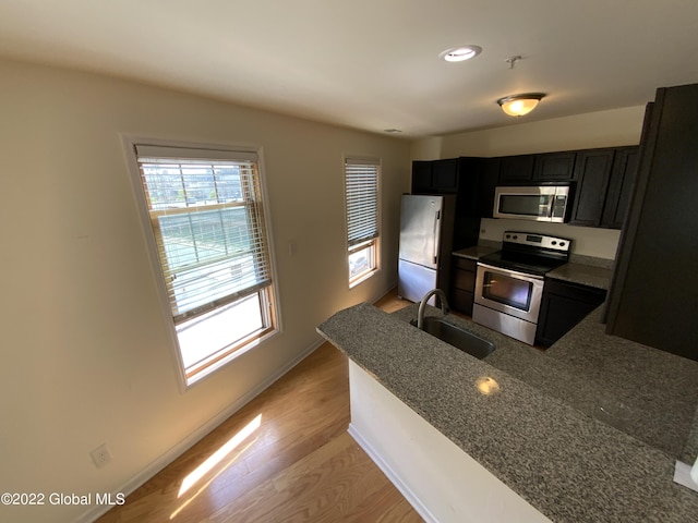 kitchen with a sink, dark cabinetry, appliances with stainless steel finishes, a peninsula, and light wood-style flooring