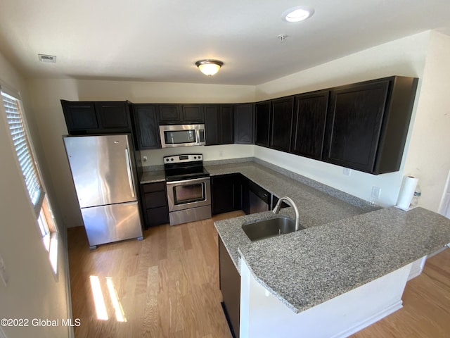 kitchen featuring a sink, a peninsula, stainless steel appliances, light wood-style floors, and dark cabinets