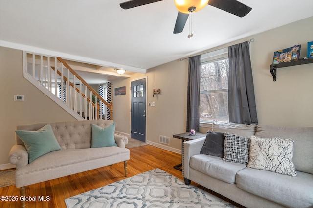 living room featuring a ceiling fan, visible vents, wood finished floors, baseboards, and stairs
