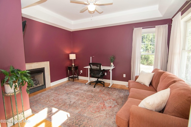 living room featuring a tray ceiling, a ceiling fan, wood finished floors, a glass covered fireplace, and baseboards