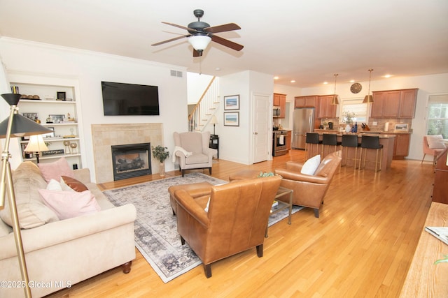 living room featuring visible vents, a tile fireplace, stairs, a ceiling fan, and light wood-style floors