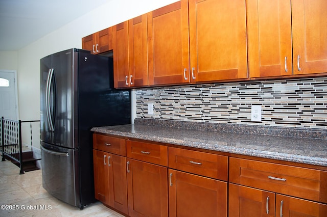 kitchen with dark stone counters, freestanding refrigerator, light tile patterned floors, brown cabinets, and backsplash