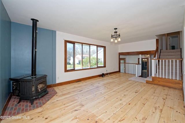 unfurnished living room featuring a notable chandelier, a wood stove, stairs, baseboards, and hardwood / wood-style flooring