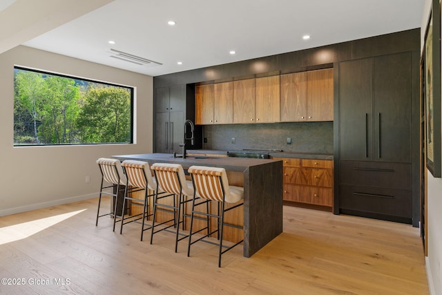 kitchen with a sink, a kitchen island with sink, dark countertops, a breakfast bar area, and light wood-type flooring