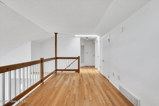 hallway with visible vents, baseboards, light wood-style floors, a textured ceiling, and an upstairs landing