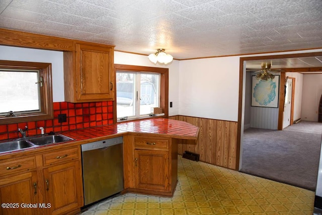 kitchen with a wainscoted wall, brown cabinetry, stainless steel dishwasher, tile countertops, and a sink