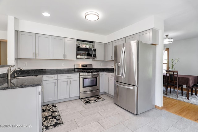 kitchen with a sink, dark stone counters, appliances with stainless steel finishes, and gray cabinetry
