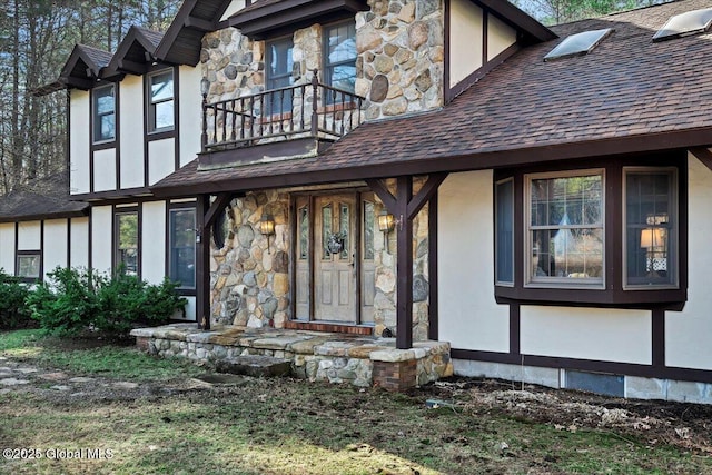 property entrance with stucco siding, roof with shingles, stone siding, and a balcony