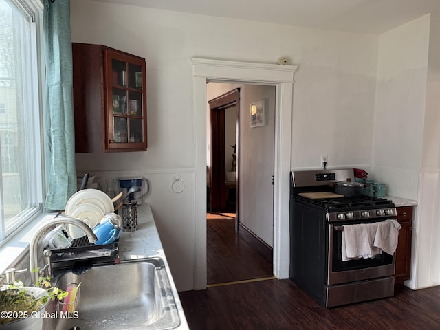 kitchen featuring a sink, stainless steel gas stove, glass insert cabinets, light countertops, and dark wood-style flooring