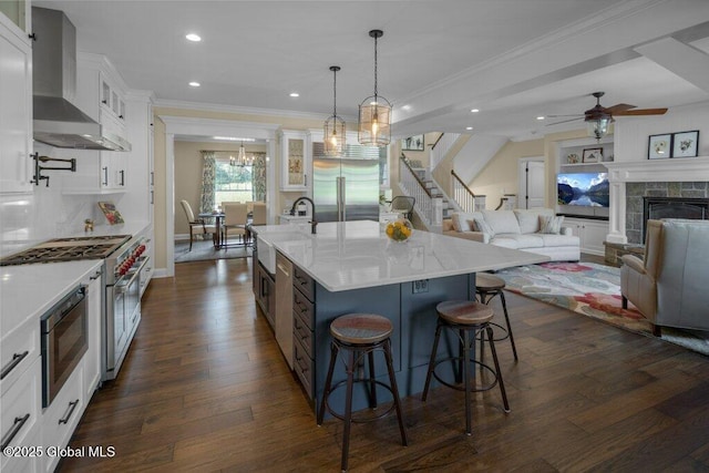 kitchen with built in appliances, wall chimney range hood, dark wood-type flooring, a sink, and ceiling fan with notable chandelier