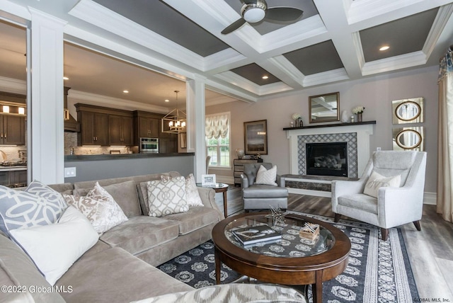 living area featuring wood finished floors, ceiling fan with notable chandelier, a tiled fireplace, coffered ceiling, and ornamental molding