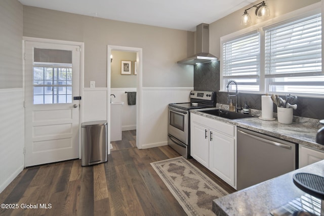 kitchen featuring appliances with stainless steel finishes, white cabinetry, a sink, a wainscoted wall, and wall chimney exhaust hood