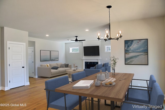 dining space featuring light wood finished floors, a premium fireplace, ceiling fan with notable chandelier, recessed lighting, and baseboards