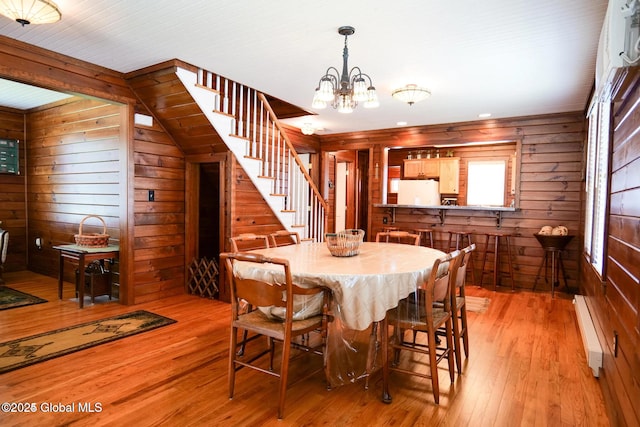 dining area featuring wood walls, a baseboard radiator, light wood finished floors, a notable chandelier, and stairs
