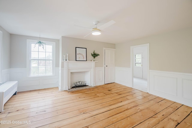 unfurnished living room featuring wainscoting, light wood-type flooring, a ceiling fan, radiator, and a decorative wall