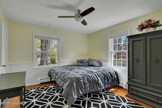 bedroom featuring a baseboard radiator, wood finished floors, a wainscoted wall, a baseboard heating unit, and ceiling fan