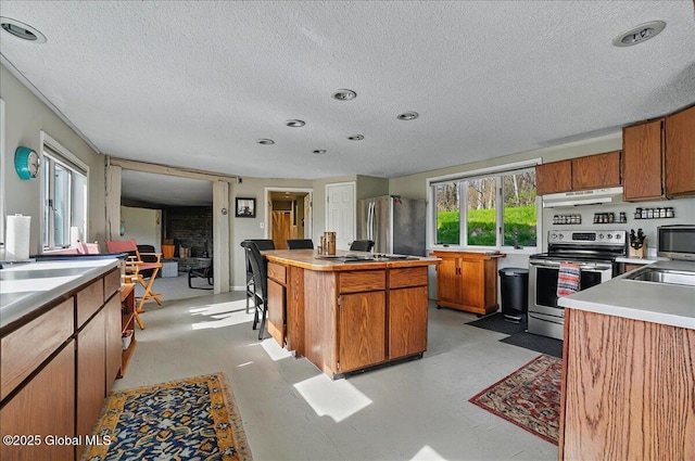 kitchen with brown cabinetry, a center island, a textured ceiling, under cabinet range hood, and appliances with stainless steel finishes