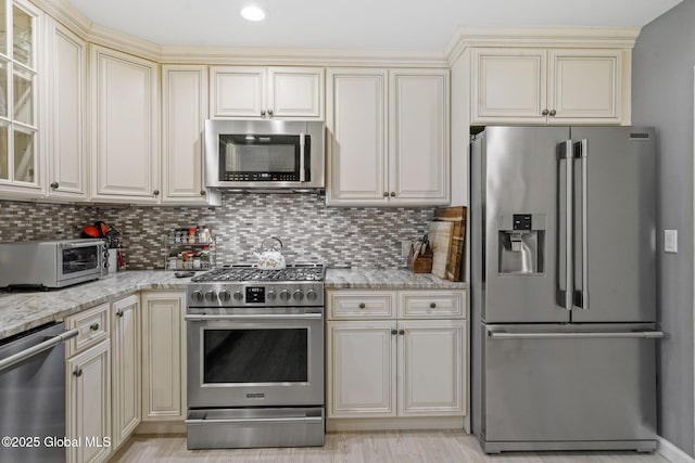 kitchen featuring stainless steel appliances, cream cabinetry, light stone countertops, and backsplash