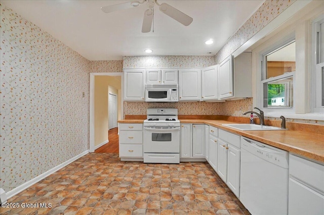 kitchen featuring white appliances, wallpapered walls, a sink, baseboards, and white cabinets