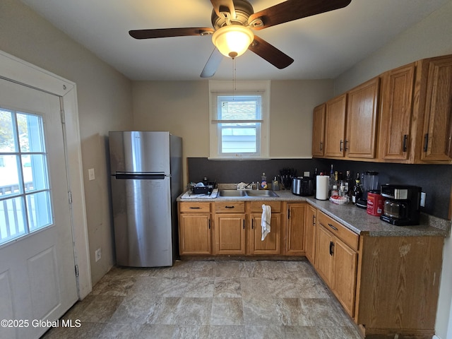 kitchen with freestanding refrigerator, a ceiling fan, stone finish floor, brown cabinets, and a sink