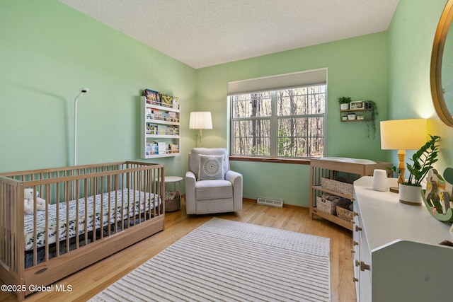 bedroom with a textured ceiling, visible vents, a nursery area, wood finished floors, and baseboards