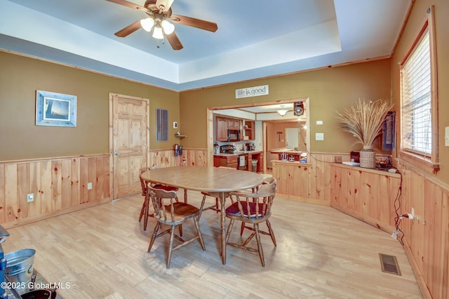 dining space with a wainscoted wall, a tray ceiling, a ceiling fan, light wood-style flooring, and wooden walls