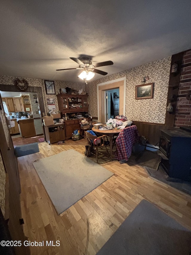 living room with ceiling fan, light wood finished floors, a wood stove, a wainscoted wall, and wallpapered walls