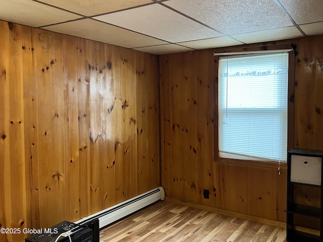 unfurnished room with a paneled ceiling, wood walls, baseboards, a baseboard heating unit, and light wood-style floors