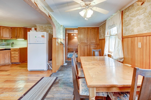 dining room with light wood-style flooring, wallpapered walls, a wainscoted wall, a ceiling fan, and arched walkways