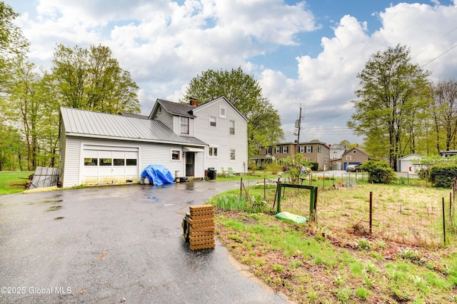view of front facade with an attached garage, asphalt driveway, a chimney, fence, and metal roof