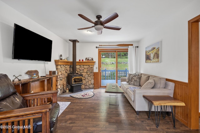 living room featuring ceiling fan, wainscoting, a wood stove, and wood finished floors
