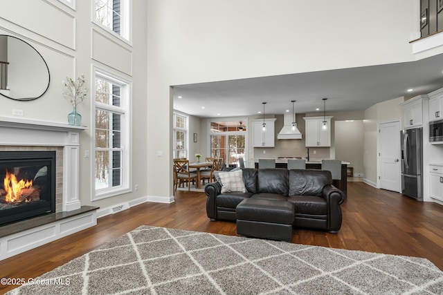 living area featuring dark wood-type flooring, a towering ceiling, a glass covered fireplace, baseboards, and recessed lighting