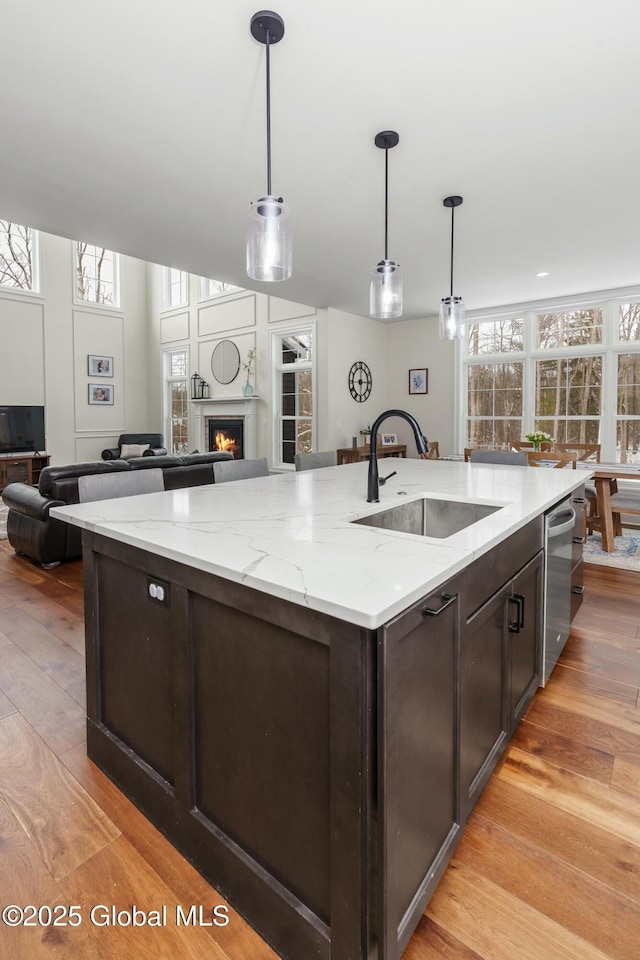 kitchen with a sink, an island with sink, light wood-style floors, dark brown cabinetry, and a lit fireplace