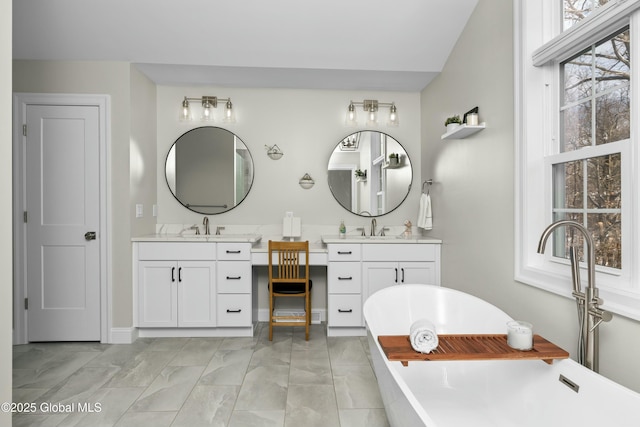 full bath with marble look tile flooring, a freestanding bath, two vanities, and baseboards